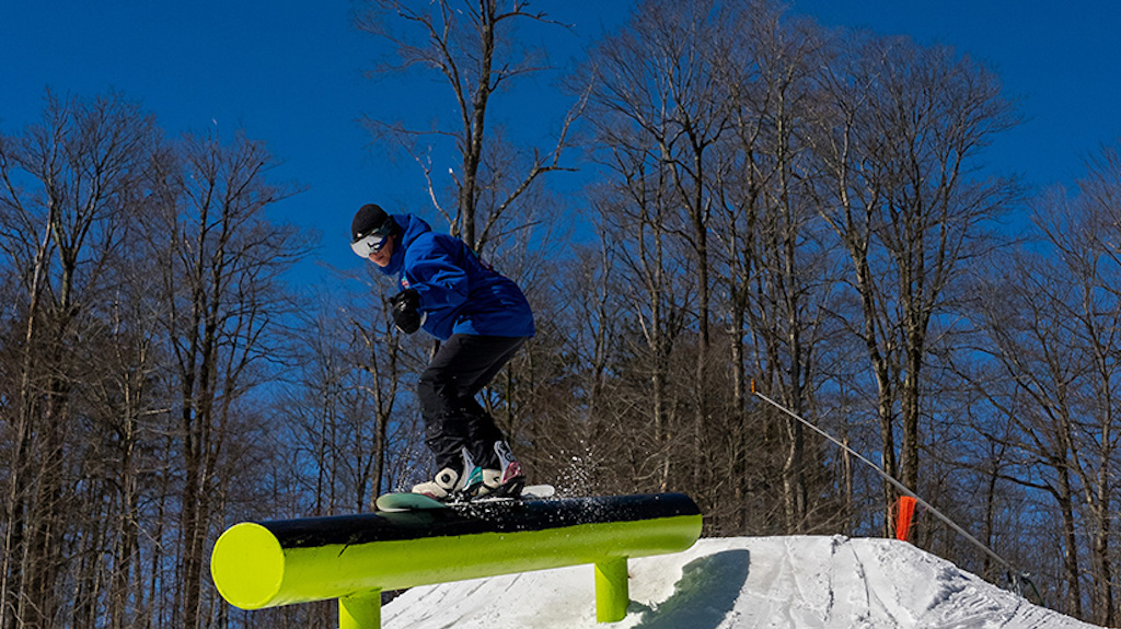 A snowboarder 50/50's a barrel rail in the park on a sunny day.